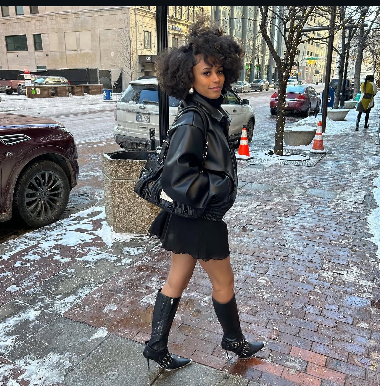 Young woman with curly hair in a black jacket and skirt, standing on a snowy city sidewalk with cars and trees.