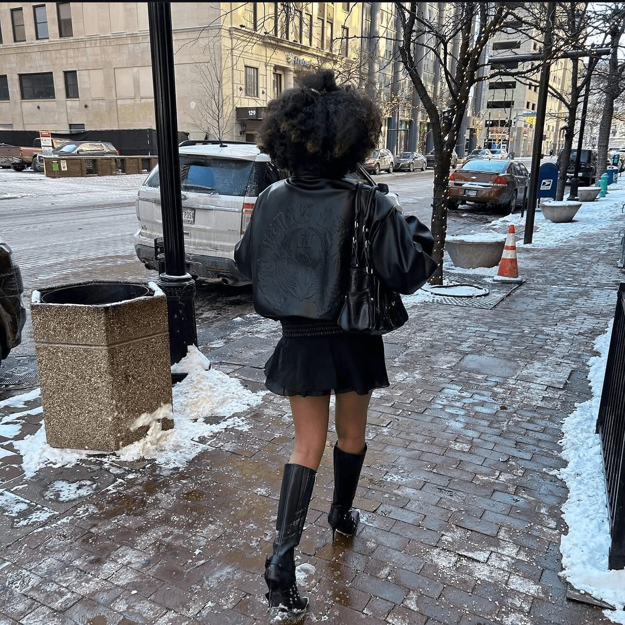 Person walking on a snowy city sidewalk with trees and parked cars, wearing a black jacket, skirt, and tall boots.