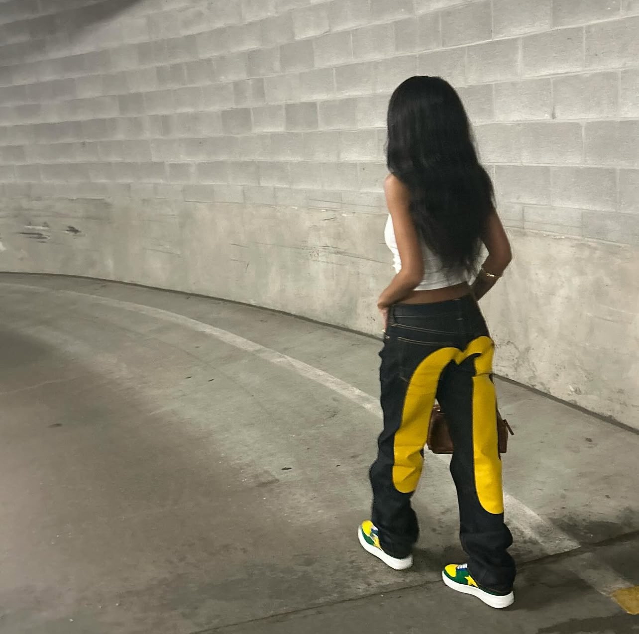 Person with long dark hair standing with back to camera in an underground parking lot, wearing black and yellow pants and sneakers.