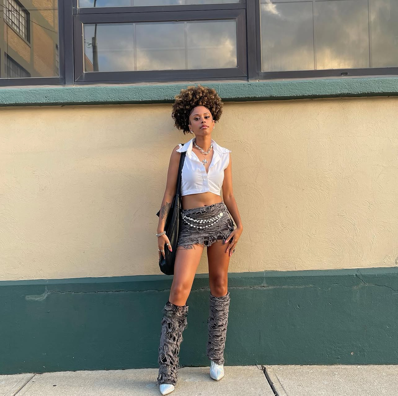 Young woman with curly hair standing against a beige and green wall, wearing a white crop top, patterned shorts, and thigh-high boots.