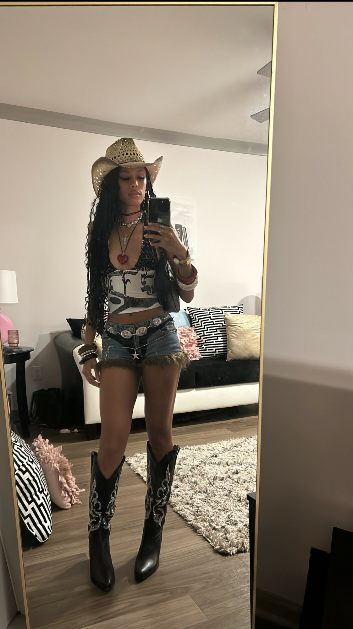 Person taking mirror selfie wearing cowboy hat, sunglasses, patterned top, denim shorts, and cowboy boots in a living room.