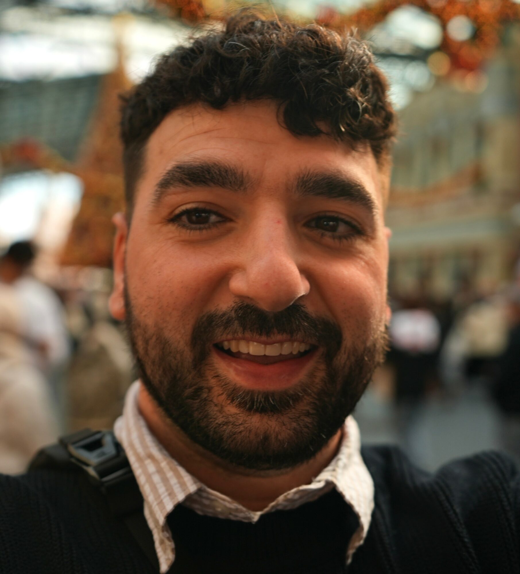 Close-up of smiling man with dark curly hair and beard, wearing a collared shirt and jacket, outdoors with blurred background.