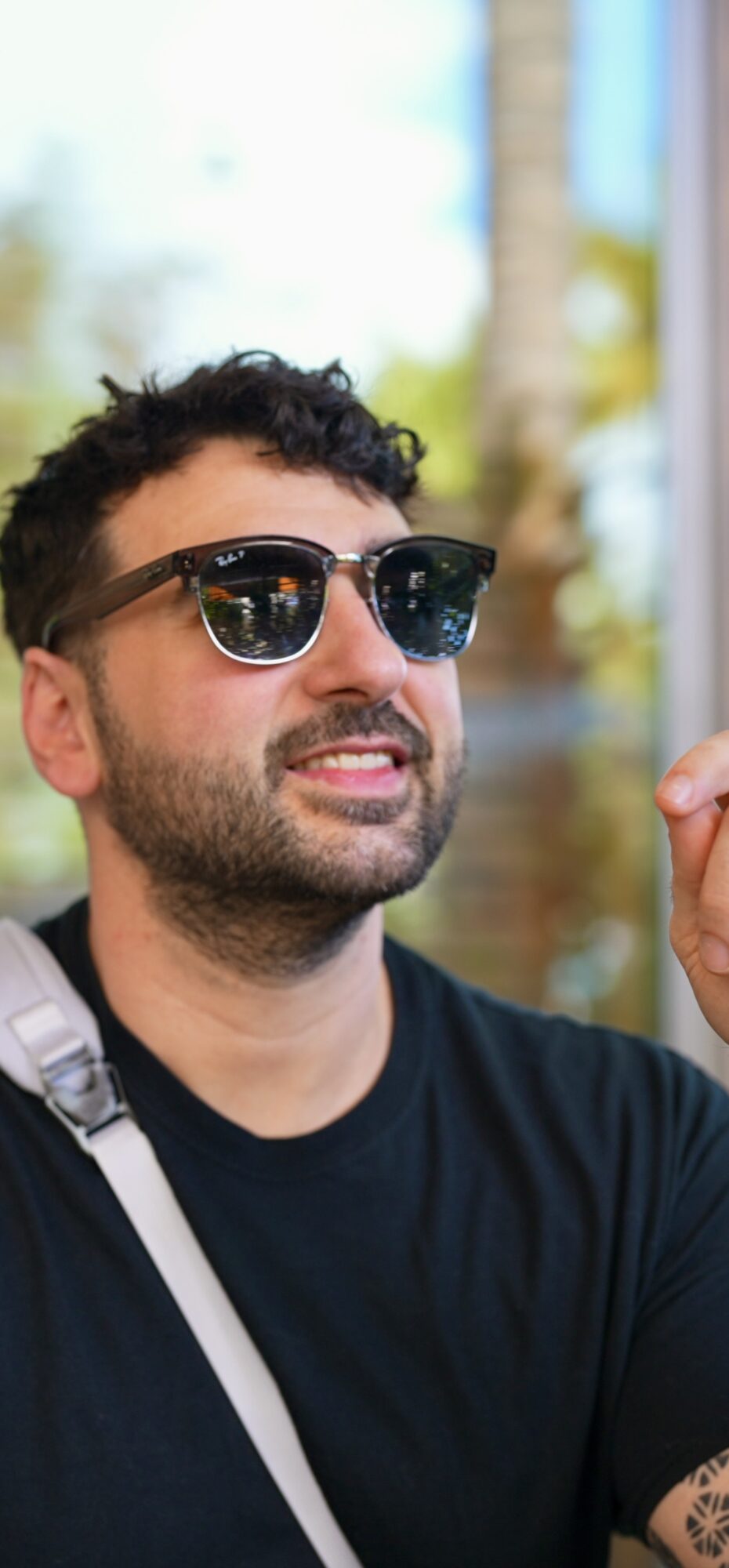 Man with dark curly hair and beard wearing sunglasses and a black shirt outdoors, holding a small object.