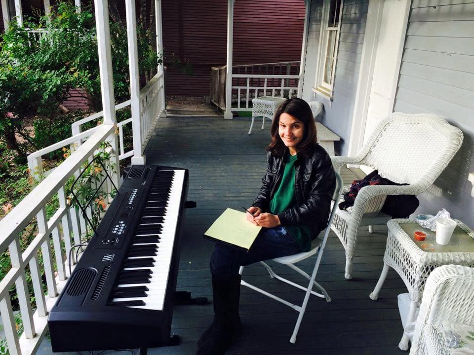 Young woman sitting on a porch with a keyboard, holding a paper, smiling, with a white chair and table nearby.
