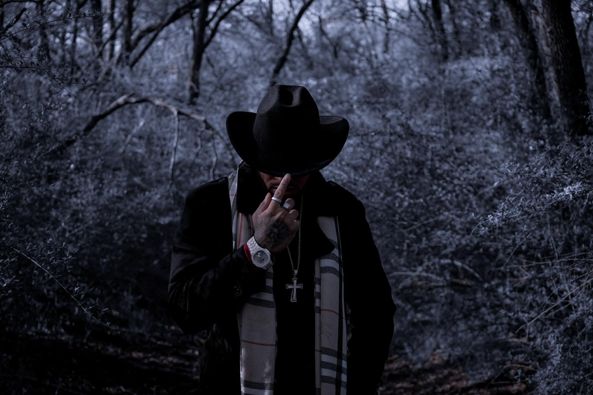 Person wearing a hat and dark clothing in a forest with snow-covered trees, holding a phone to their face.