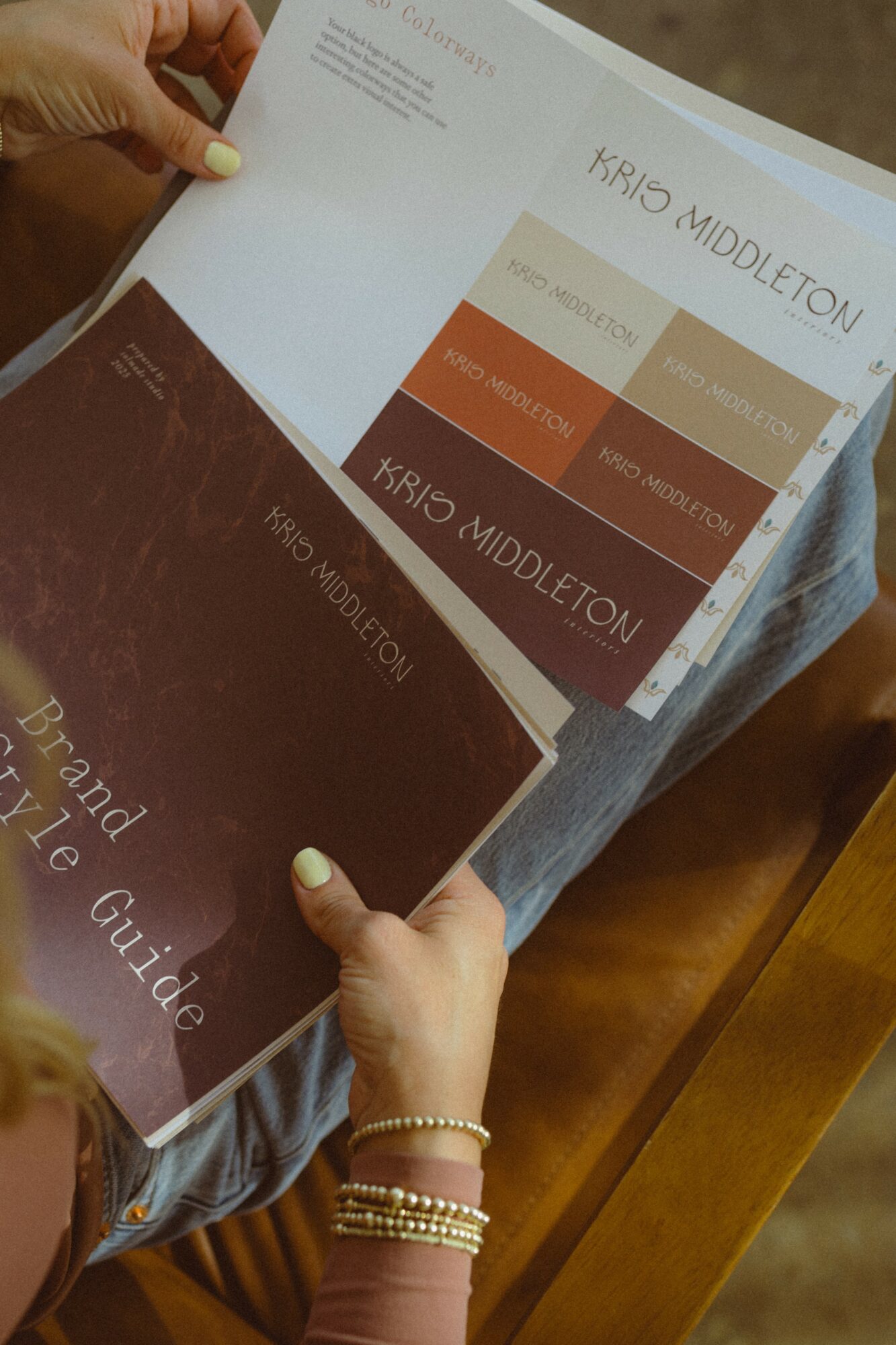 Person holding a brochure and a booklet, sitting at a wooden table, with a hand wearing bracelets. The brochure has color blocks and text.