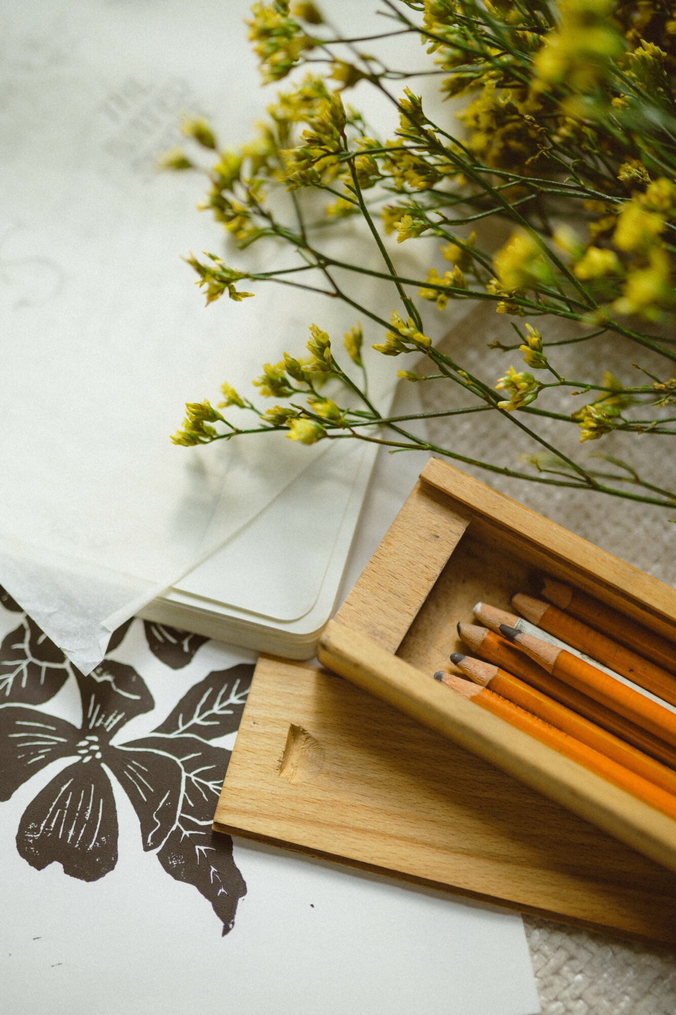 Yellow flowers with green stems, a wooden box with pencils, and a black and white leaf pattern on paper.