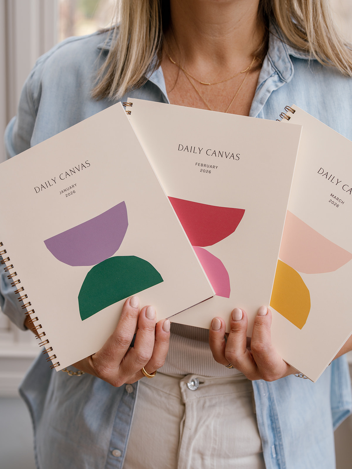 Person holding three notebooks with colorful abstract shapes on covers, wearing a light blue shirt and beige pants.