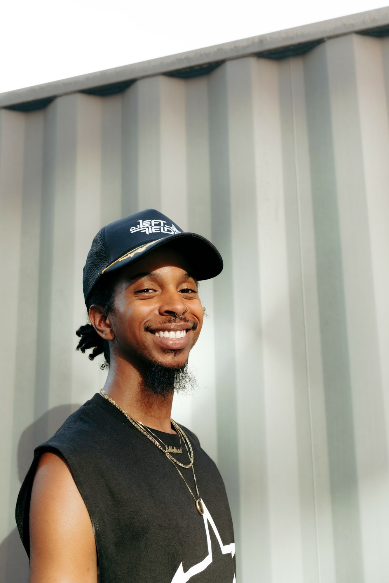 Young man smiling, wearing a black cap and sleeveless shirt, standing outdoors against a corrugated metal wall.