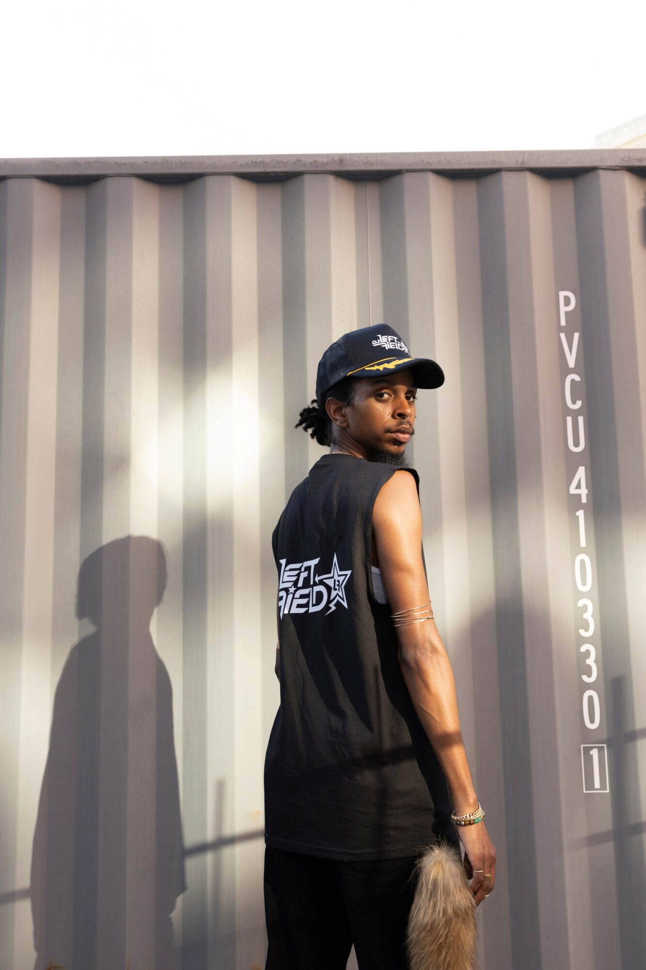 Young man with a cap and sleeveless shirt standing next to a shipping container, shadow cast on the container wall.