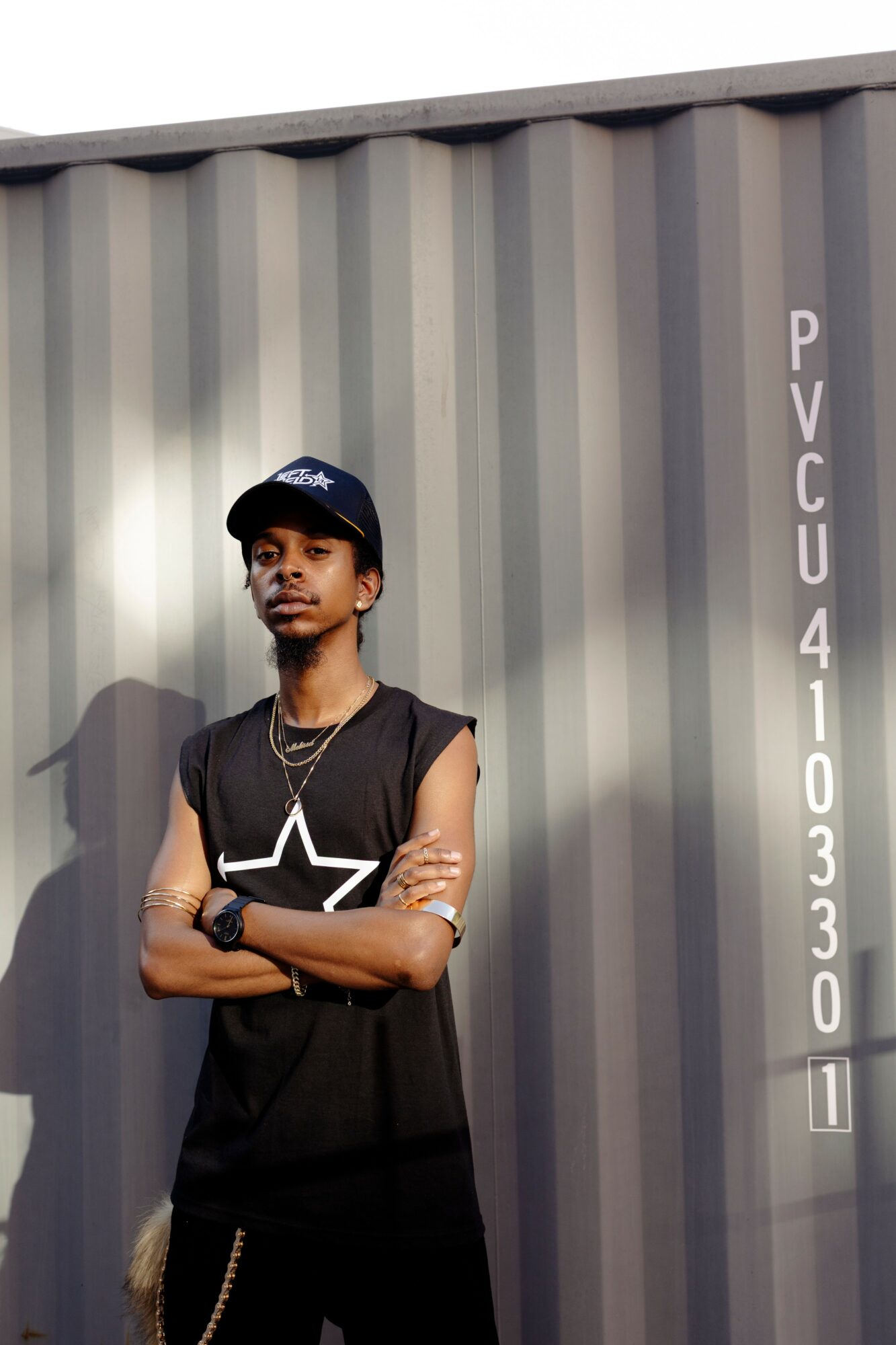Young man with arms crossed, wearing a black sleeveless shirt, cap, and jewelry, standing in front of a gray container.