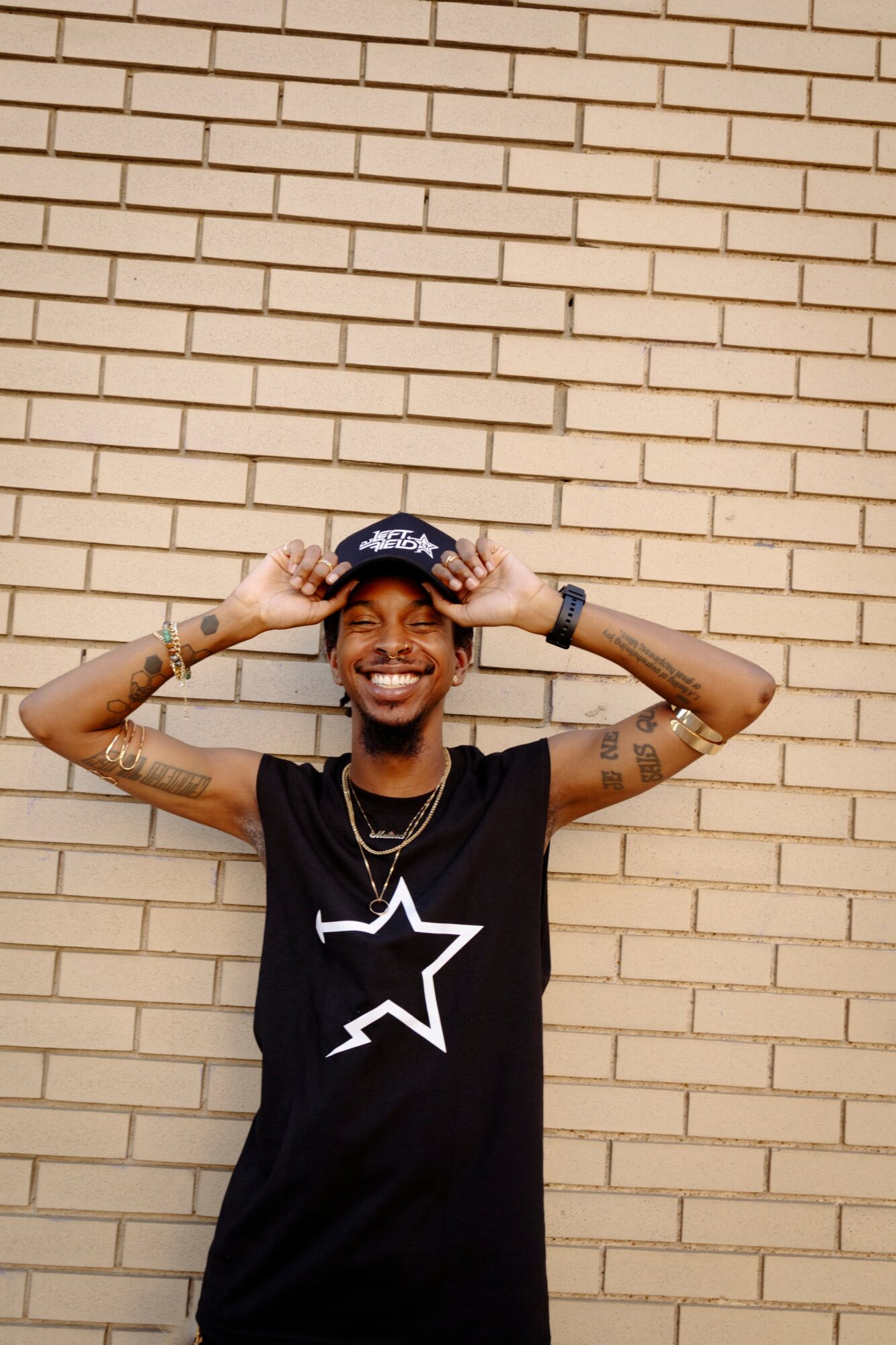 Smiling person with tattoos, wearing a black sleeveless shirt with a star logo, standing against a brick wall.