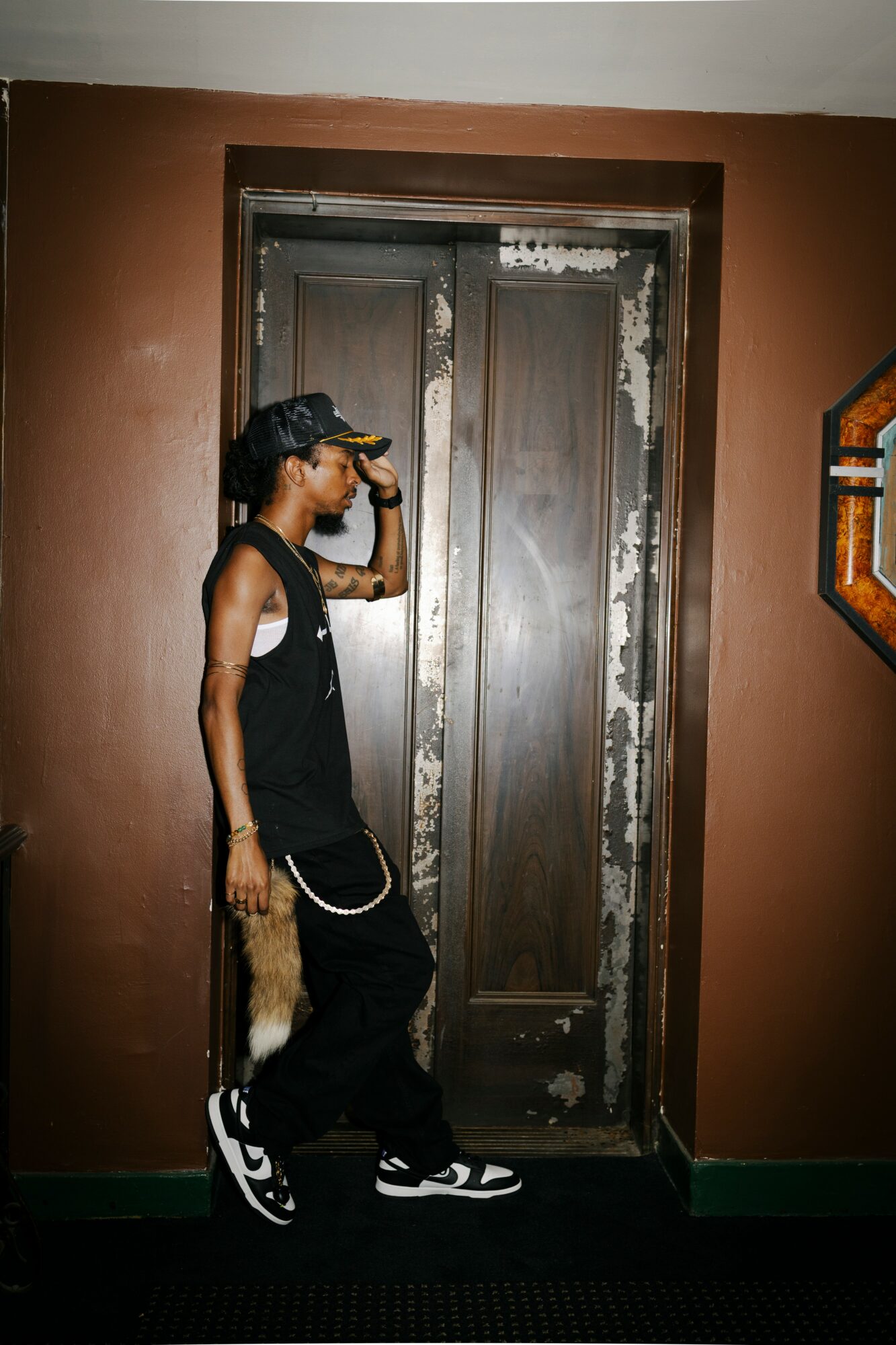 Person in black clothing and sneakers standing in front of a closed wooden door, holding a yellow object to their forehead.
