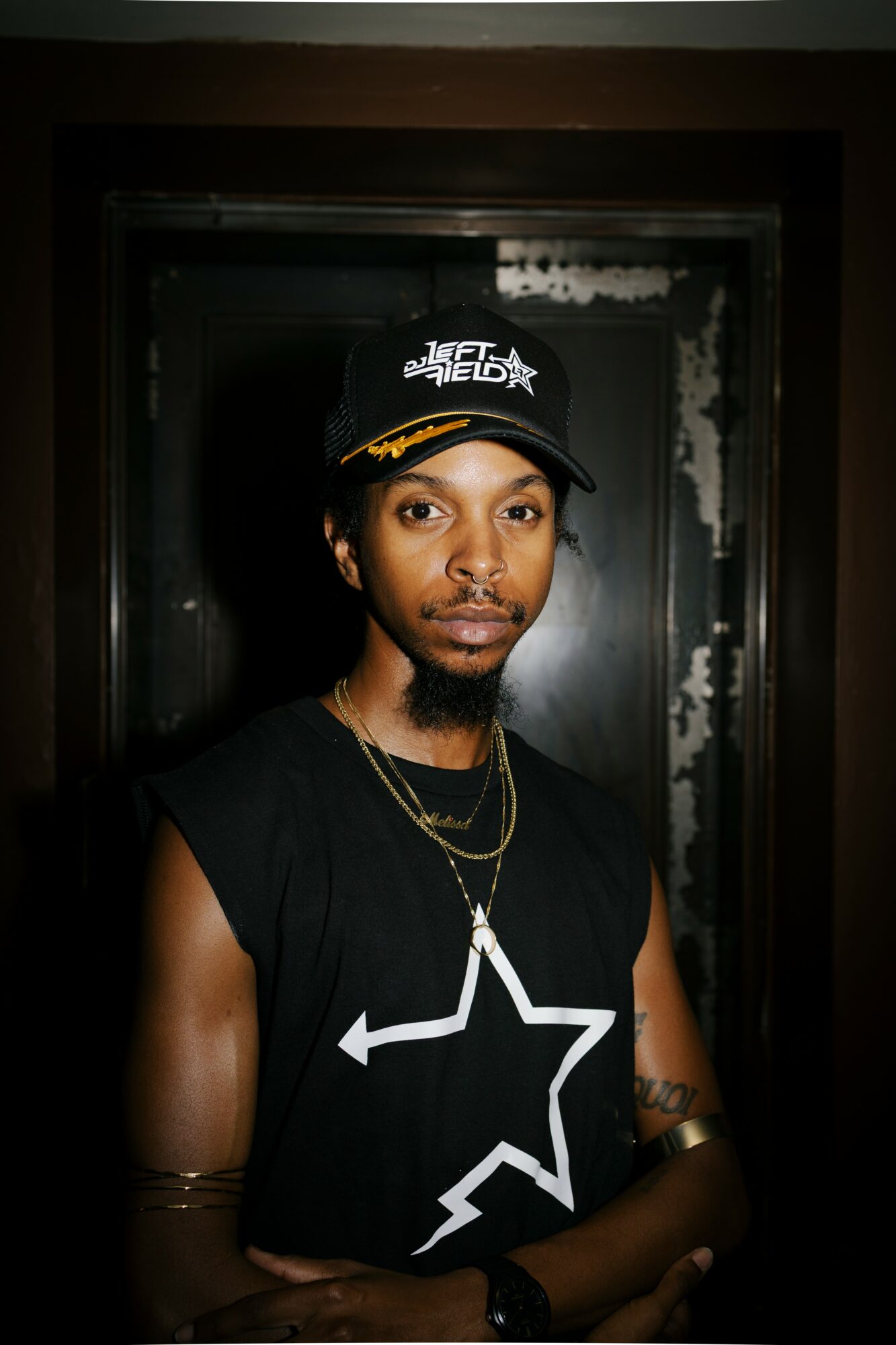 Young person wearing a black cap and sleeveless shirt, standing in front of a dark background with a doorway.