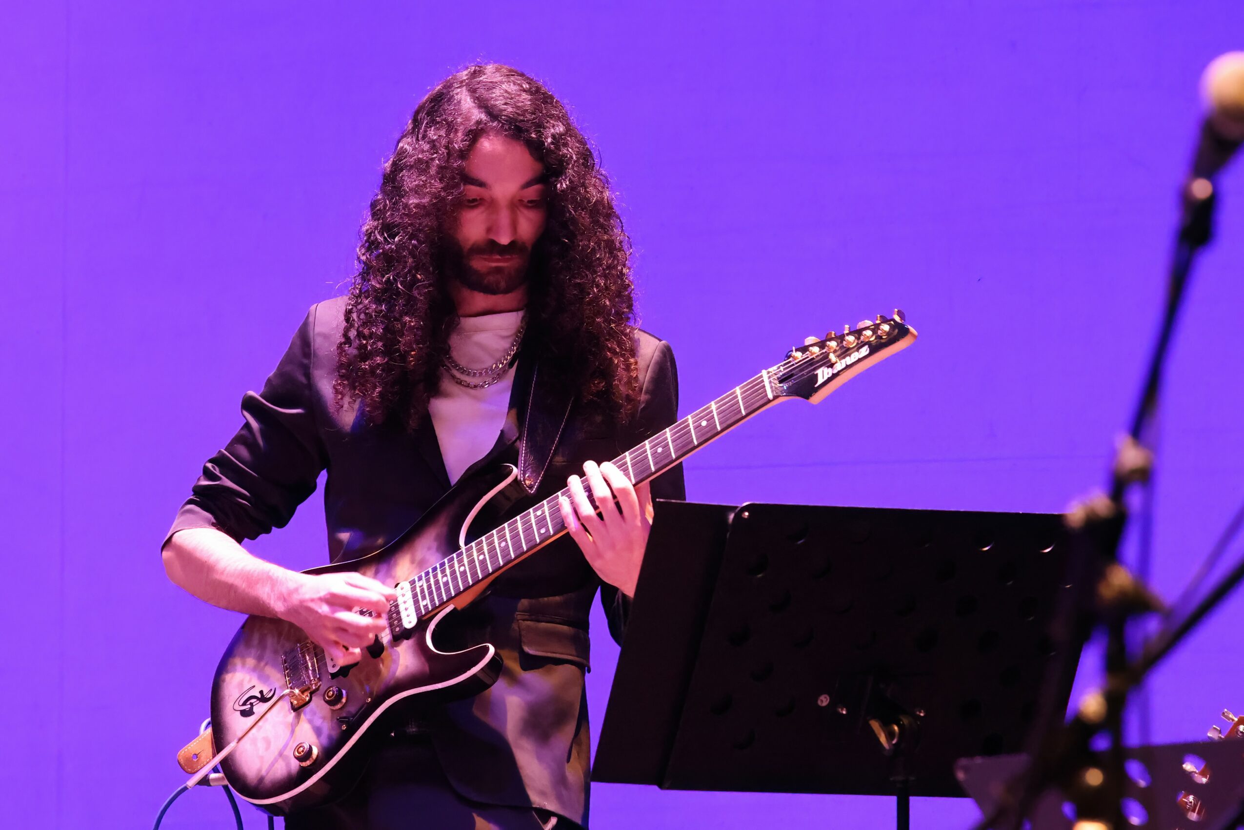 Person with long curly hair playing an electric guitar on stage with a purple background.
