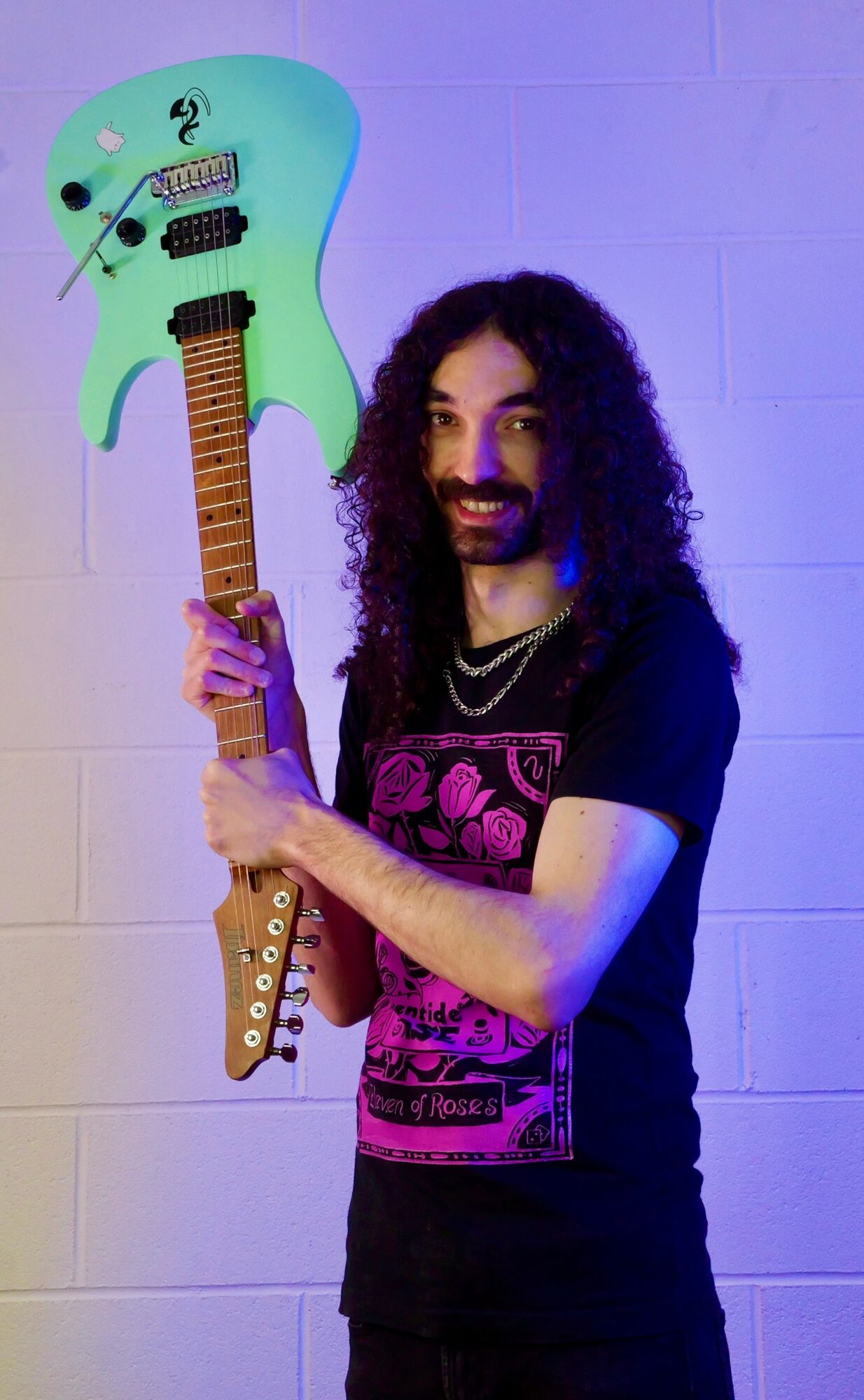 Person with long curly hair holding a colorful guitar-shaped object, smiling, standing against a light-colored wall.