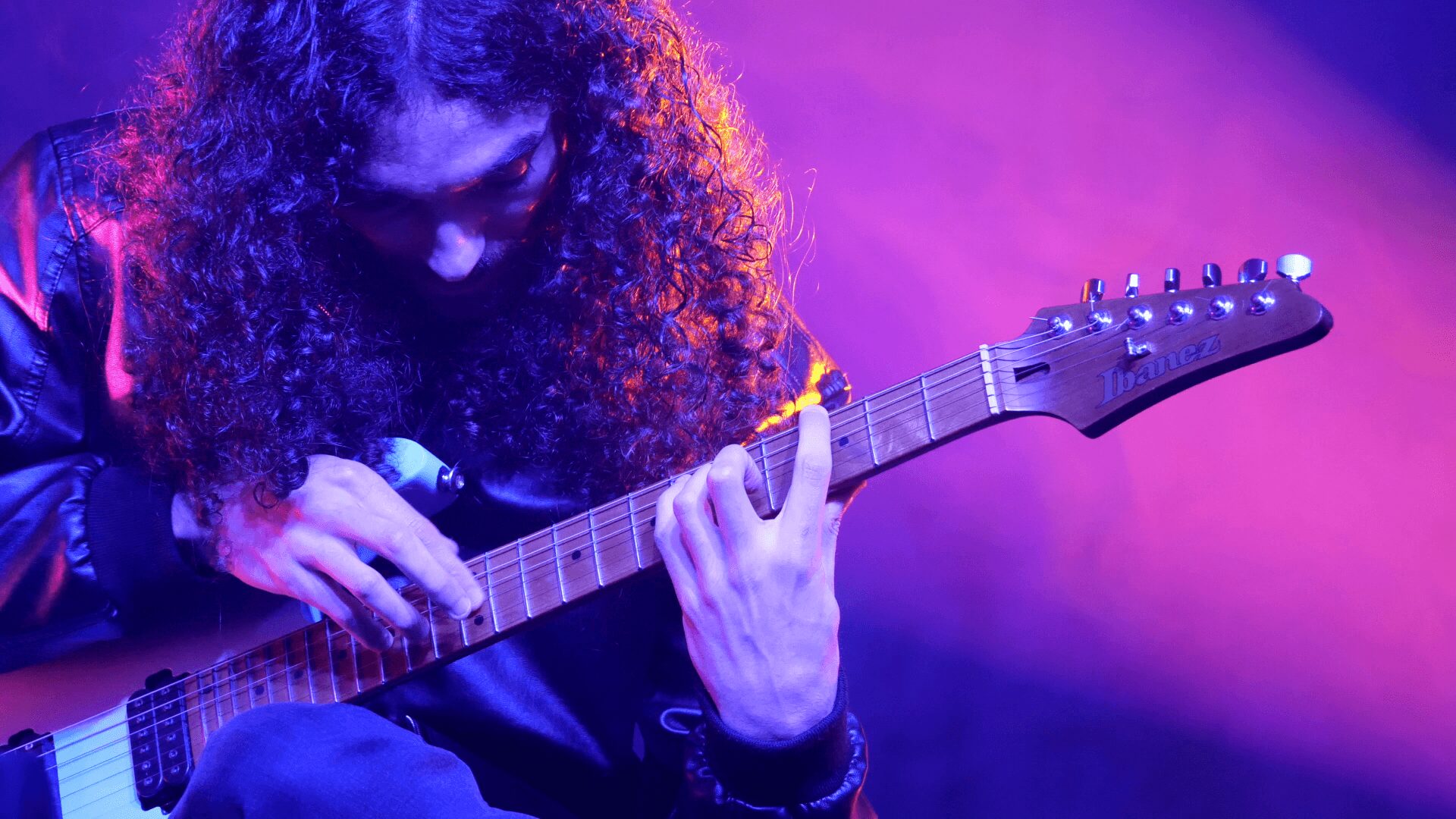 Person with long curly hair playing an electric guitar on stage with purple and pink lighting.