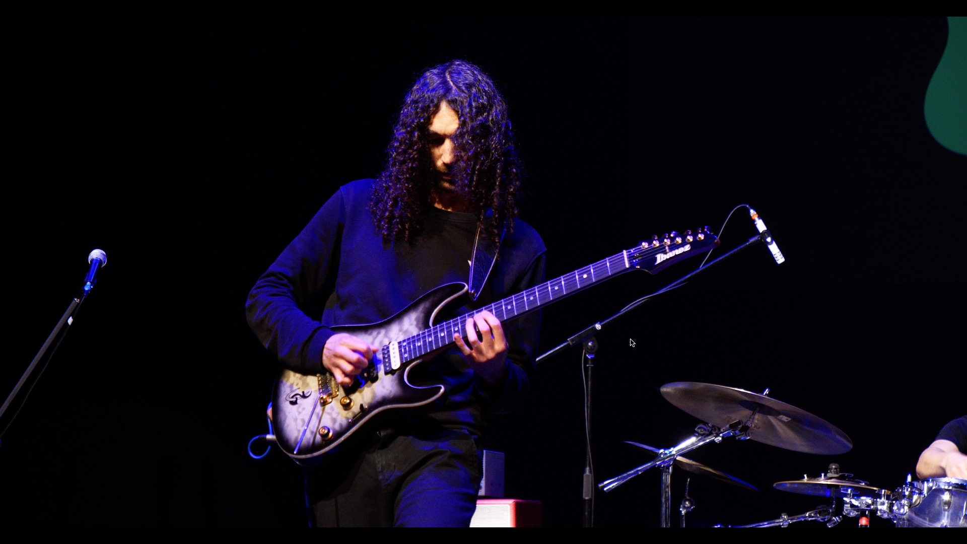Person with curly hair playing an electric guitar on stage, with a drum set nearby.