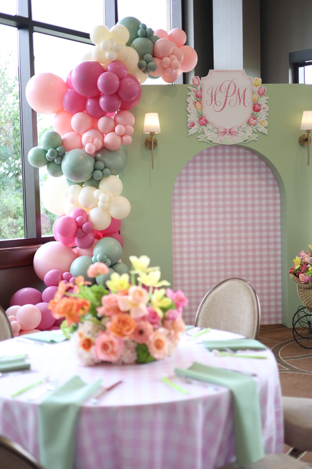 Decorative pastel balloon arrangement with flowers and a pink checkered backdrop for celebration, near a window.