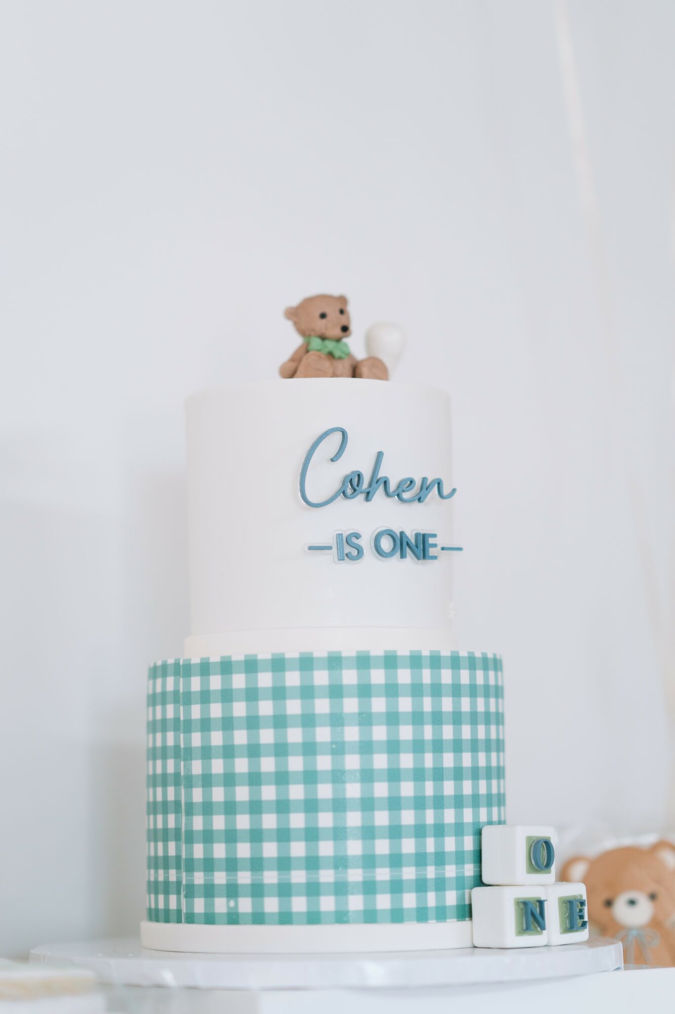 Two-tiered cake with teddy bear topper, blue checkered pattern on lower tier, and birthday message for Cohen, celebrating one year.