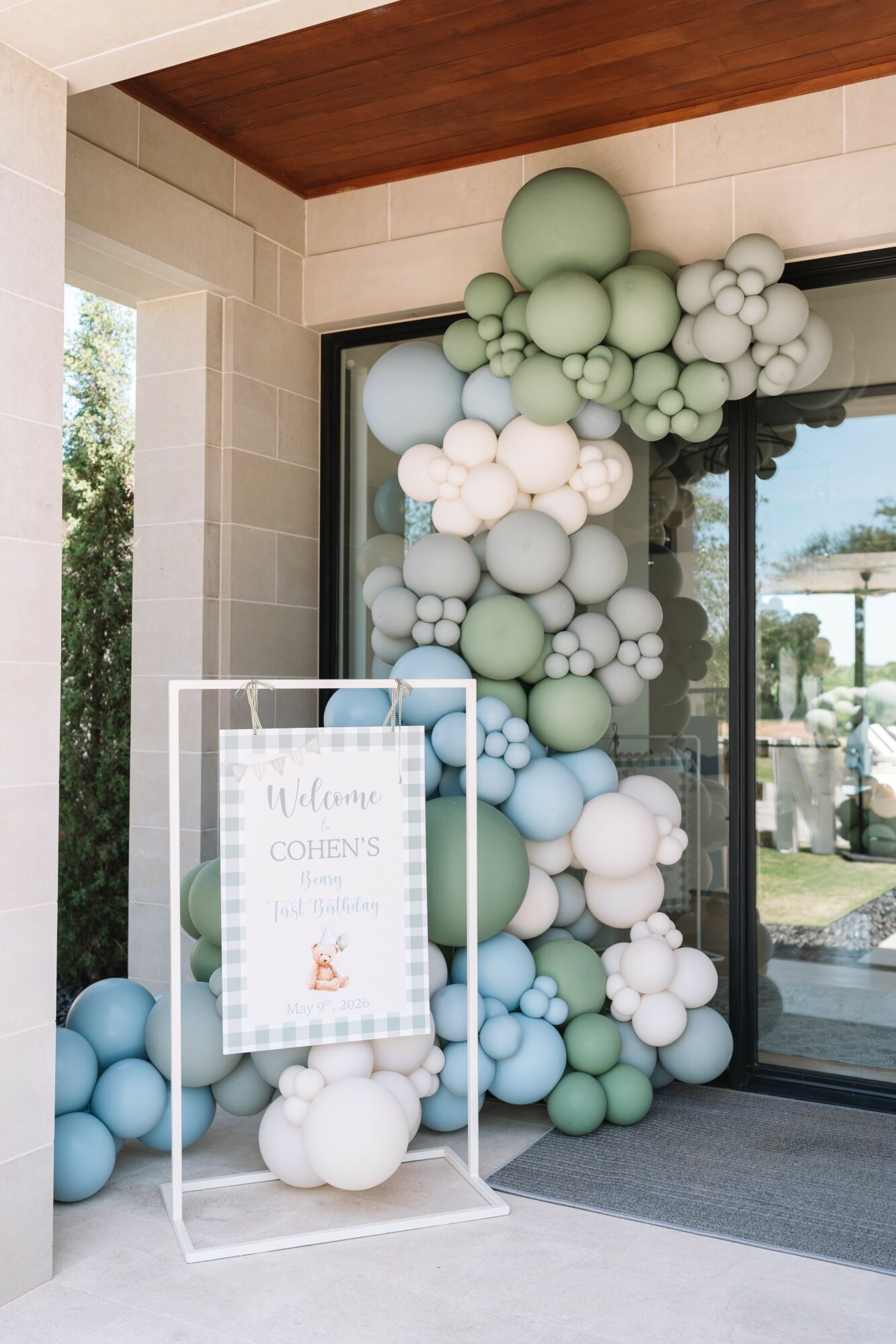 Balloon arrangement with white, light blue, and green balloons near a window and a welcome sign for a wedding.