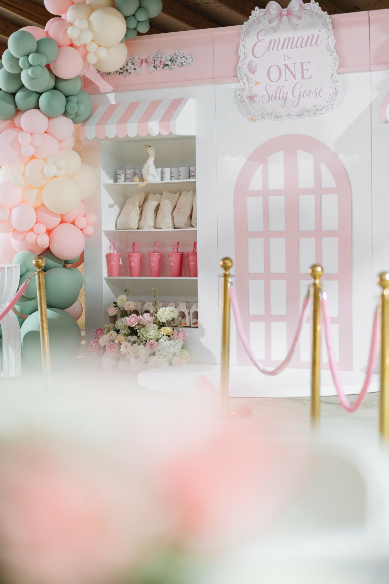 Decorative pink and white party setup with balloons, a pink door, and a display shelf with pink cups and treats.