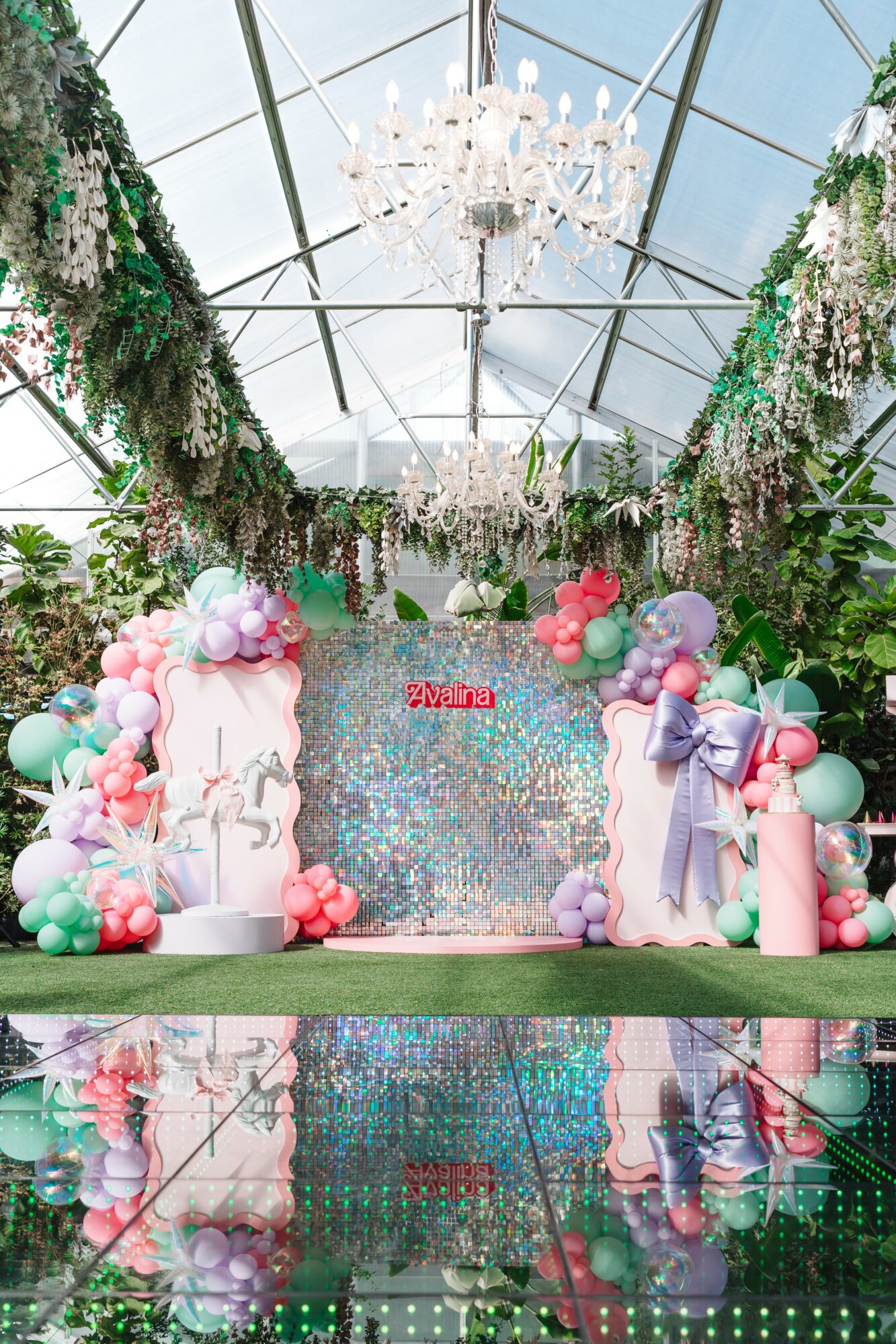 Decorative event setup with balloons, floral arrangements, and a reflective surface, under a glass ceiling with chandeliers.