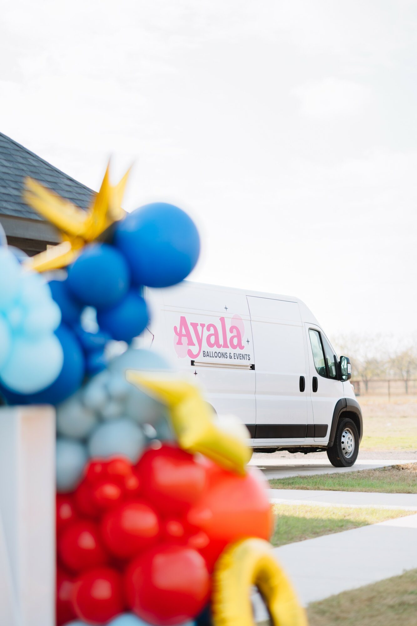 Colorful balloons in foreground with a white van labeled 'Ayala' in background, outdoors on a cloudy day.