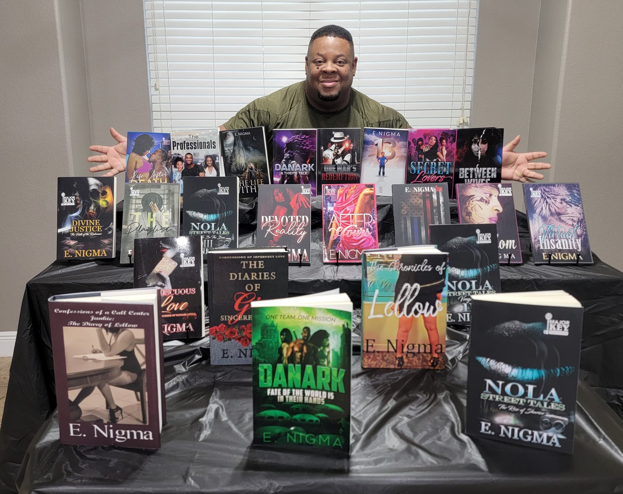 Man with arms outstretched behind a table displaying multiple books, in a room with closed blinds.