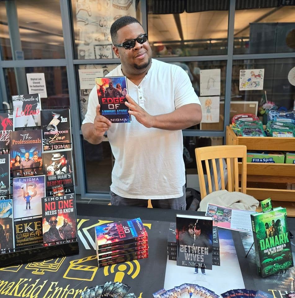 Man holding a book at a table with books and promotional materials, outside a store or cafe, smiling and wearing sunglasses.