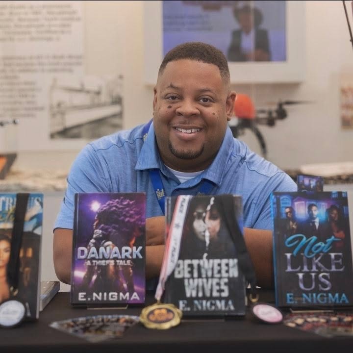 Smiling man at a table with books and medals, in a room with posters and displays in the background.