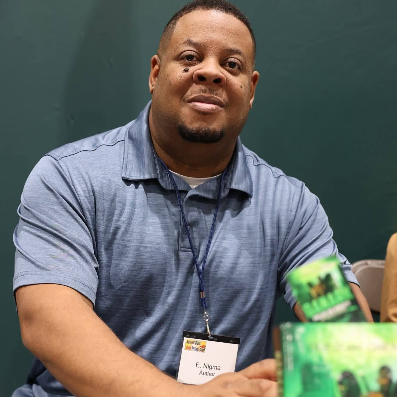 Man with short hair and goatee wearing a blue polo shirt, sitting at a table with a name tag, against a green wall.