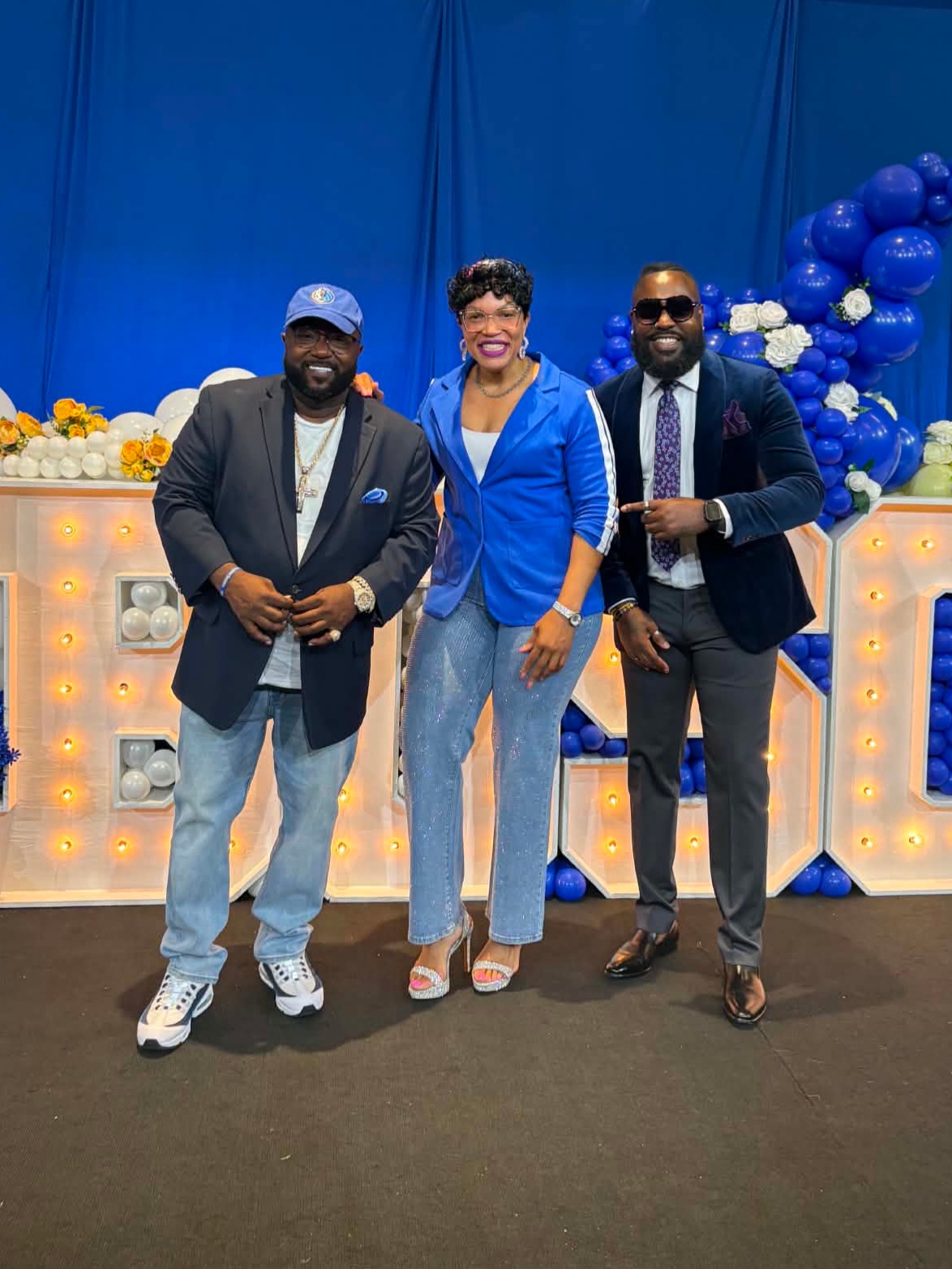 Three people standing together in front of decorated backdrop with balloons, smiling, dressed in formal and semi-formal attire.