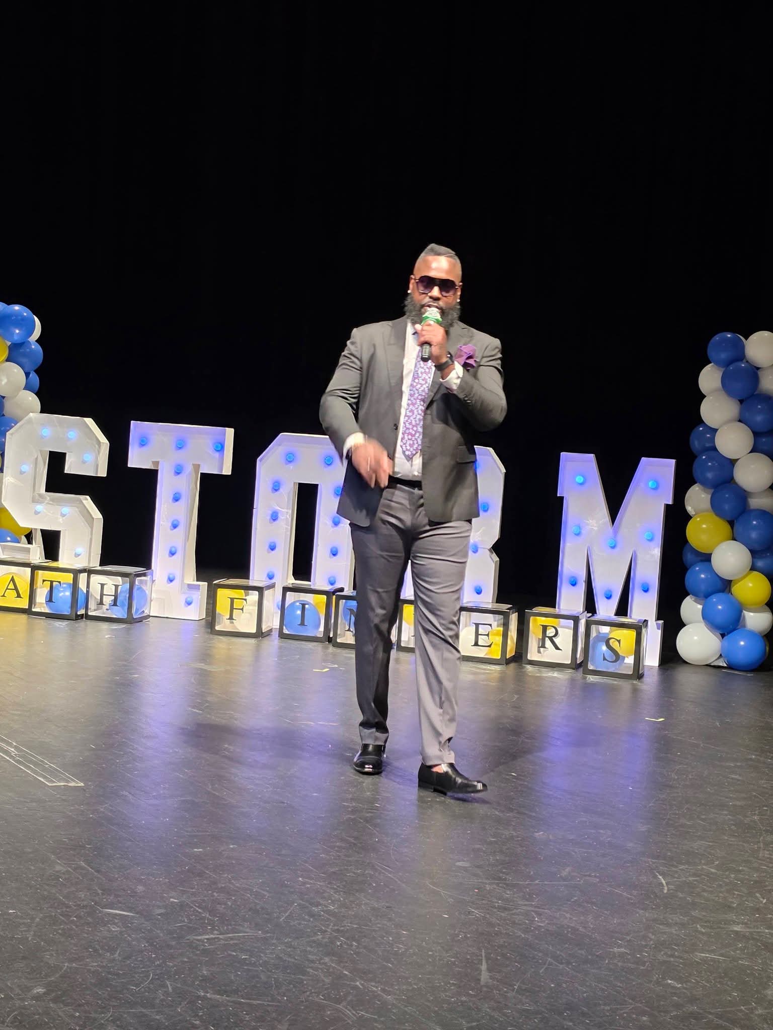 Man in suit taking a selfie on stage with large illuminated letters and balloon decorations behind him.
