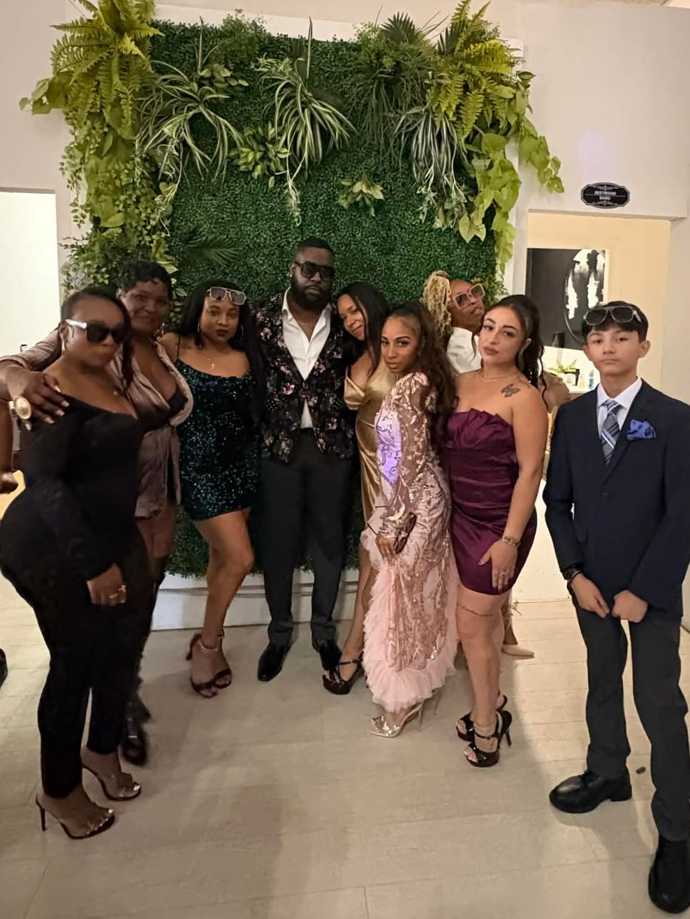 Group of nine people dressed in formal attire standing indoors in front of a green wall with plants.