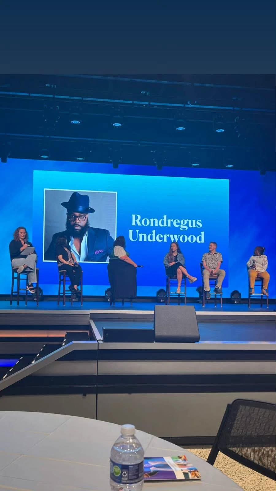 Panel discussion with five people on stage, large screen behind showing a person with a hat, blue background.