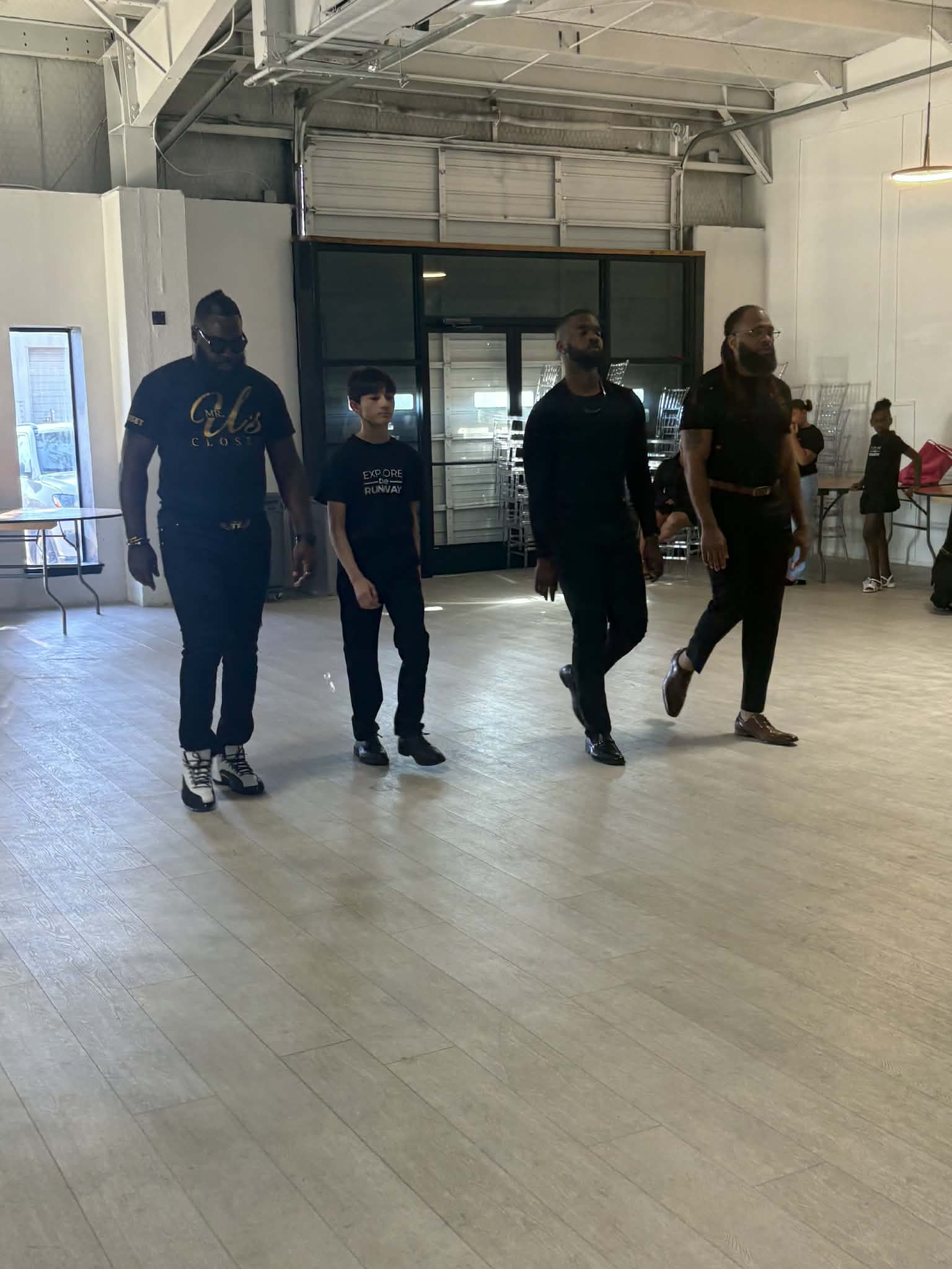 Four people walking in an indoor space with a high ceiling and large glass doors, two adults and two children, all wearing dark clothing.