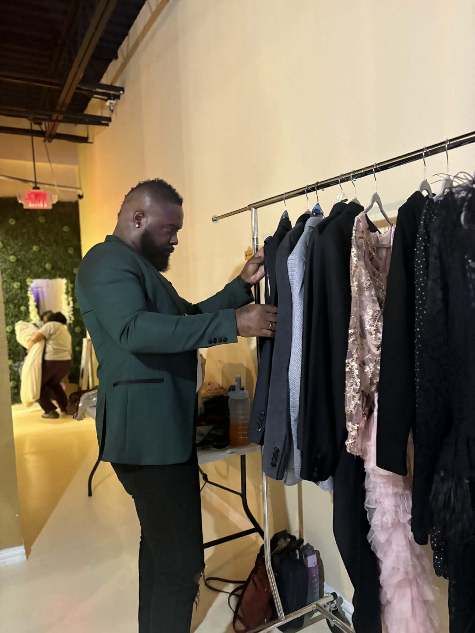 Man in green blazer browsing clothing rack with various garments in an indoor space.