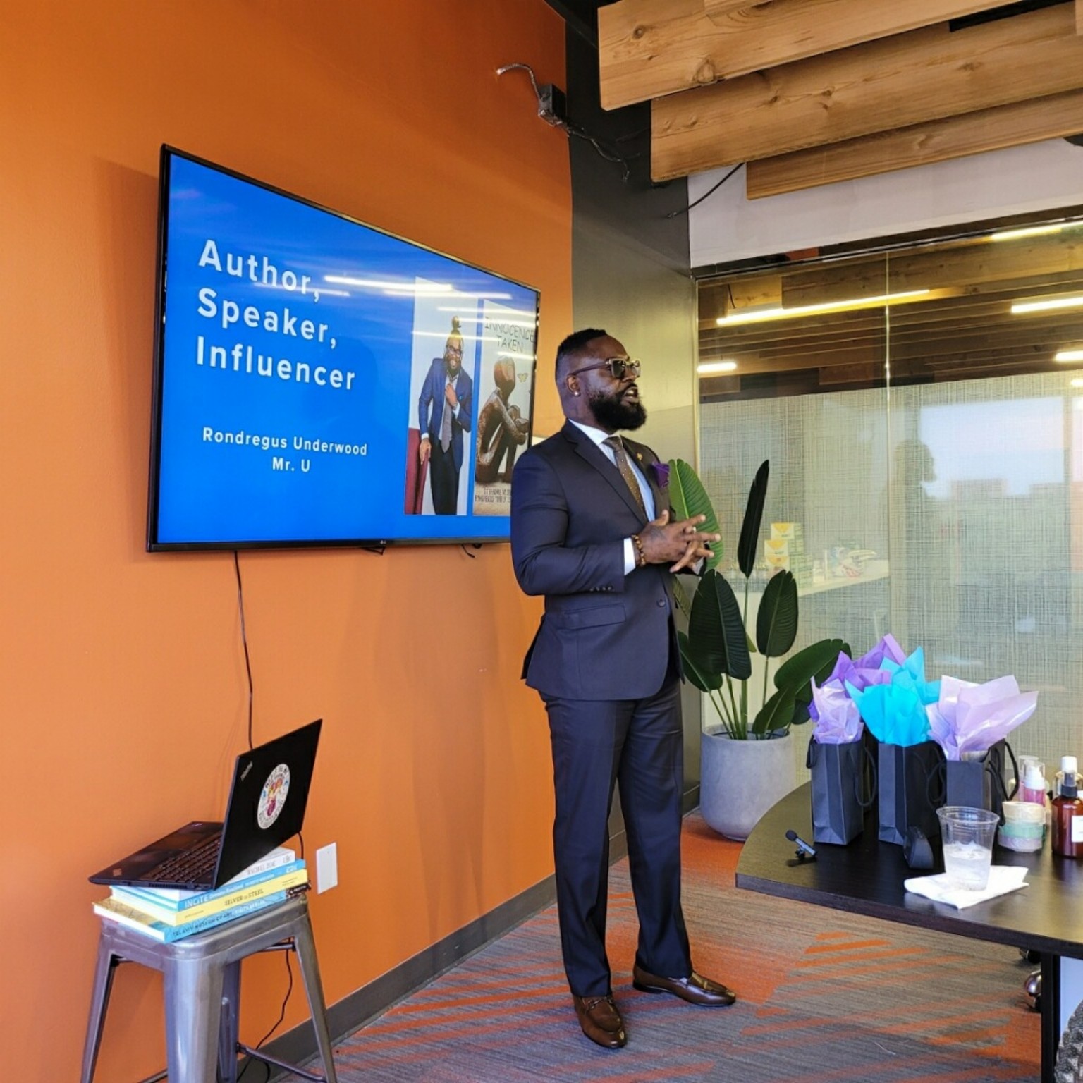Man in suit speaking in a room with orange wall, large window, and potted plant, during a presentation or event.