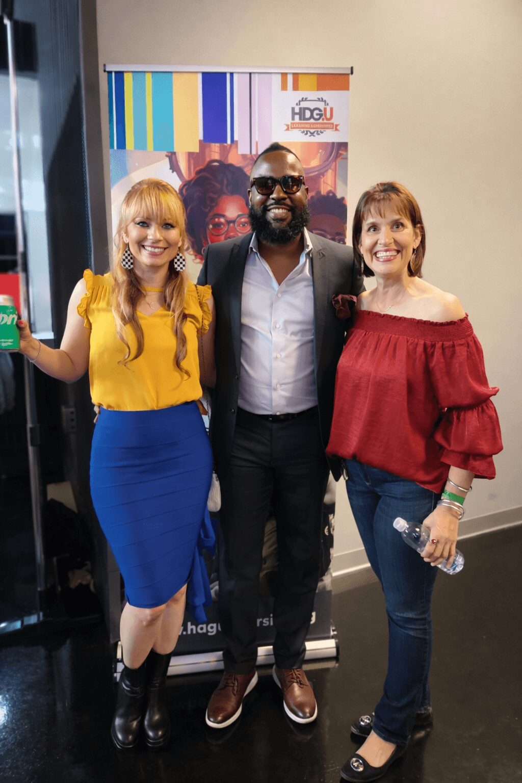 Three people standing together, smiling, in front of a colorful banner, with two women on either side of a man, all dressed casually.