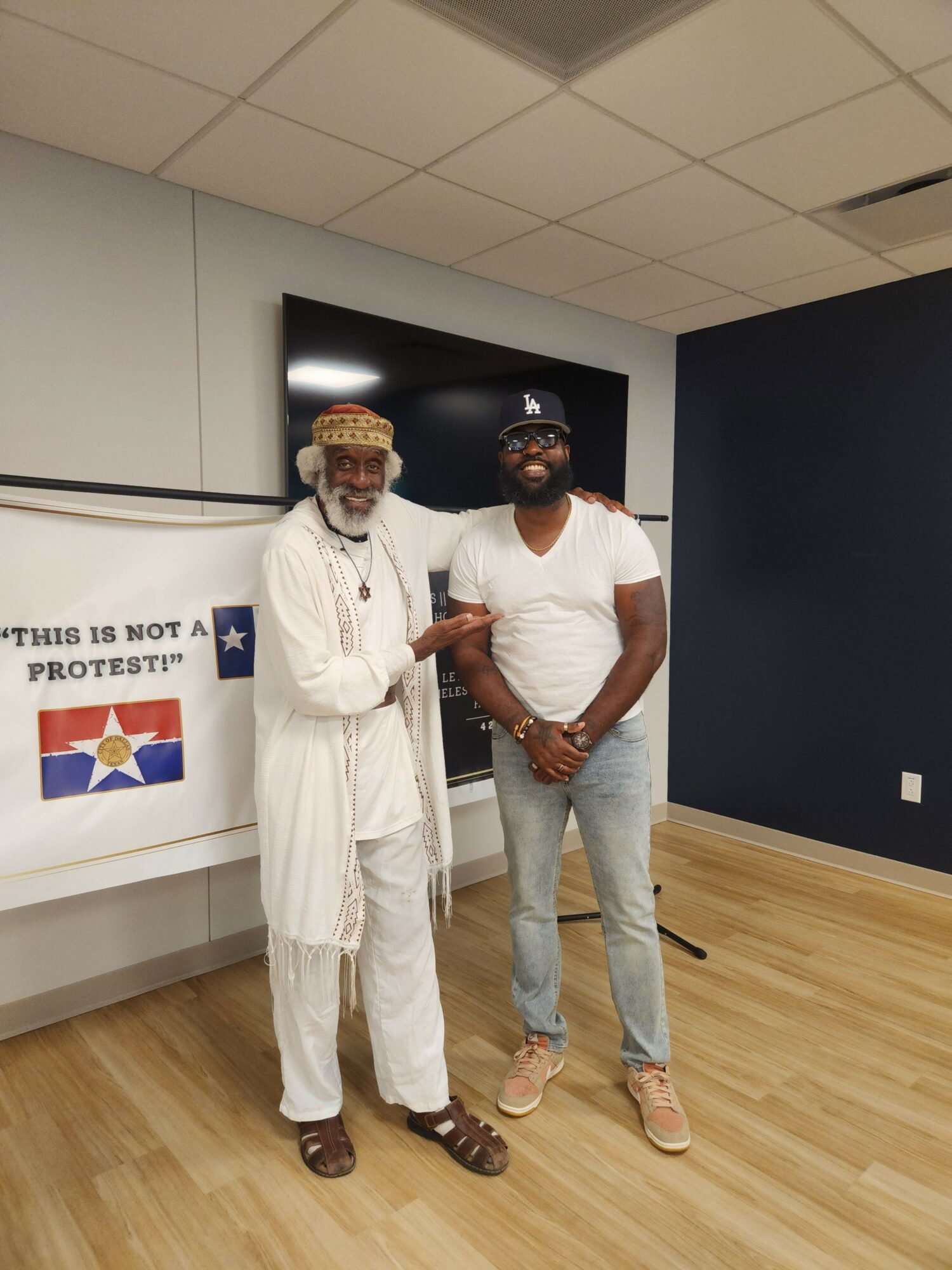 Two men standing indoors, one in casual clothes and the other in a white suit, in front of a screen and a banner.