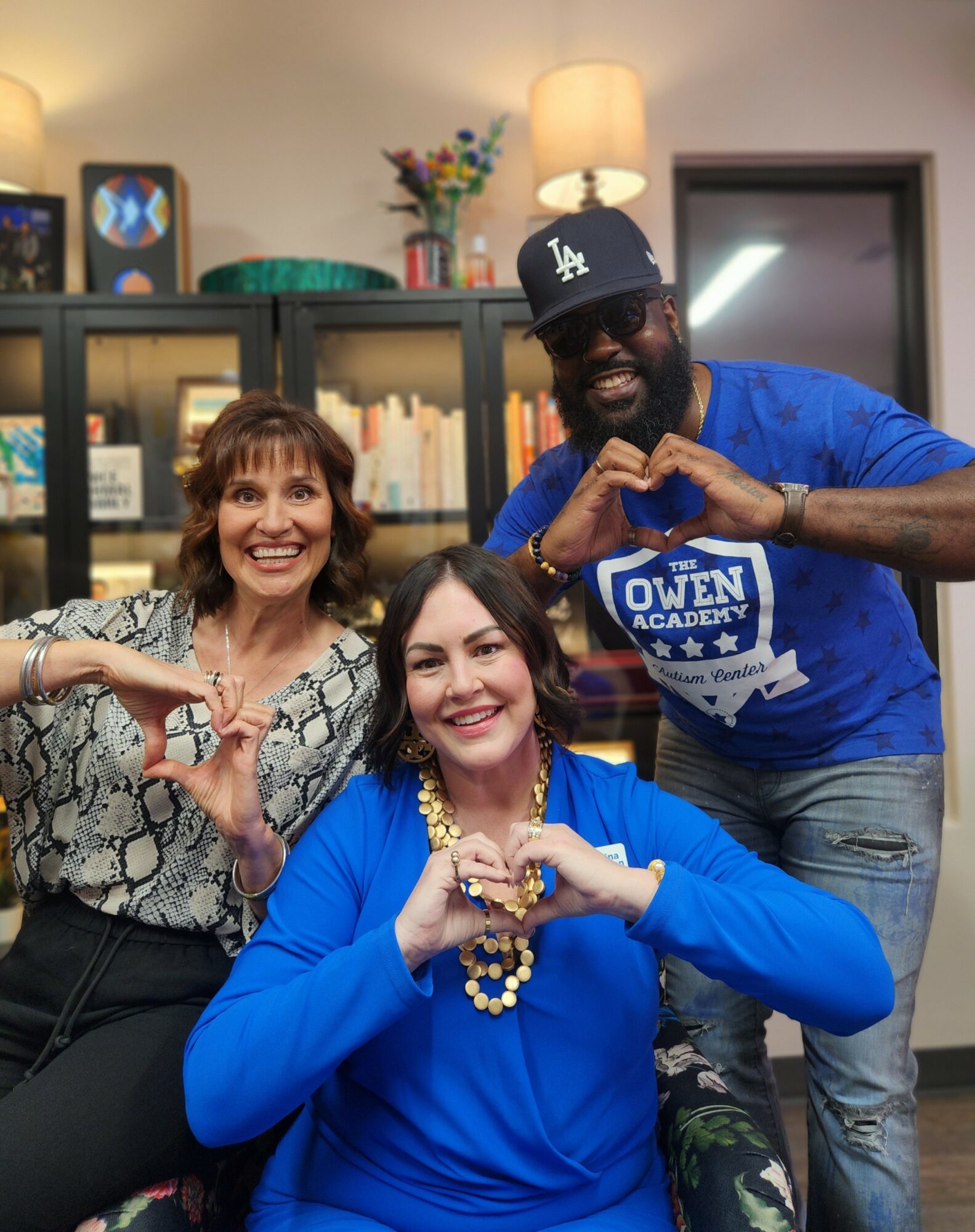 Three people smiling indoors, making heart shapes with their hands, in front of a bookshelf and lamp.