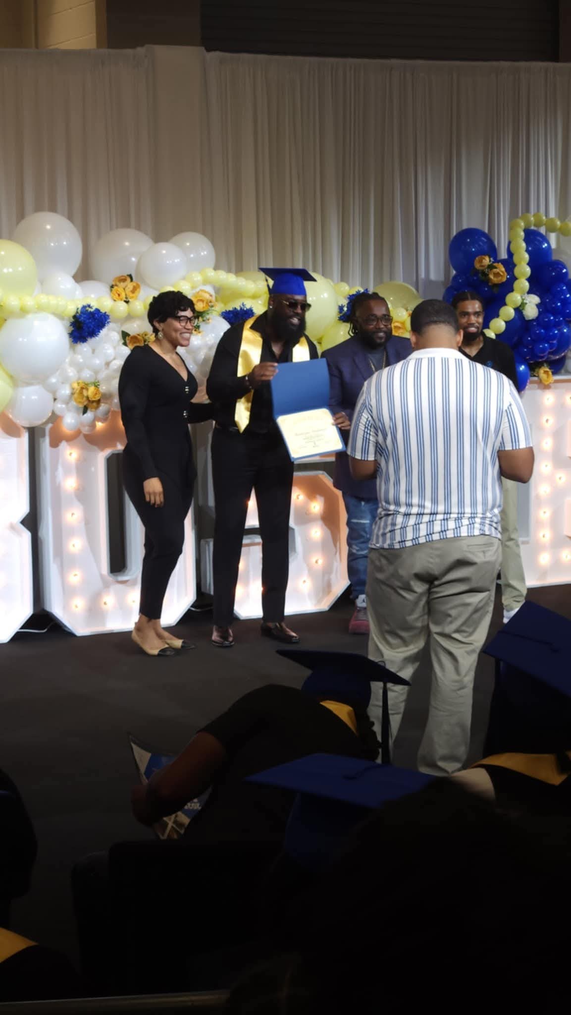 Group of people on stage with balloons and decorations, some wearing caps, in a graduation or celebration event.