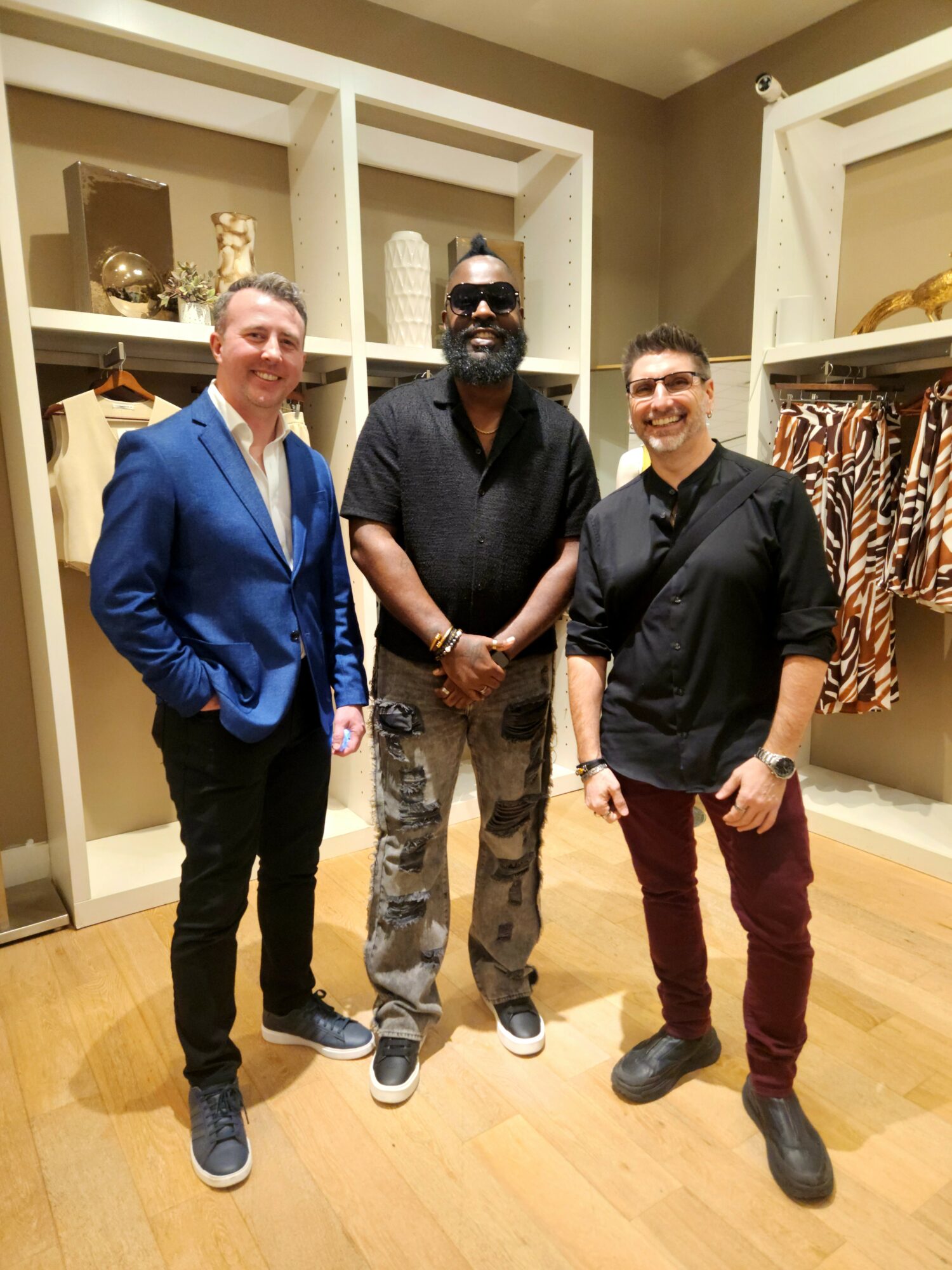 Three men standing in a clothing store, smiling, with shelves of clothes and accessories behind them.