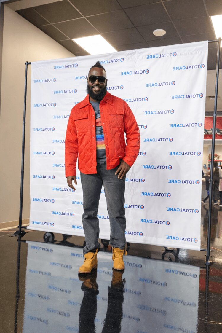 Man with sunglasses and beard wearing a red jacket, blue shirt, gray pants, and tan boots standing in front of a branded backdrop.