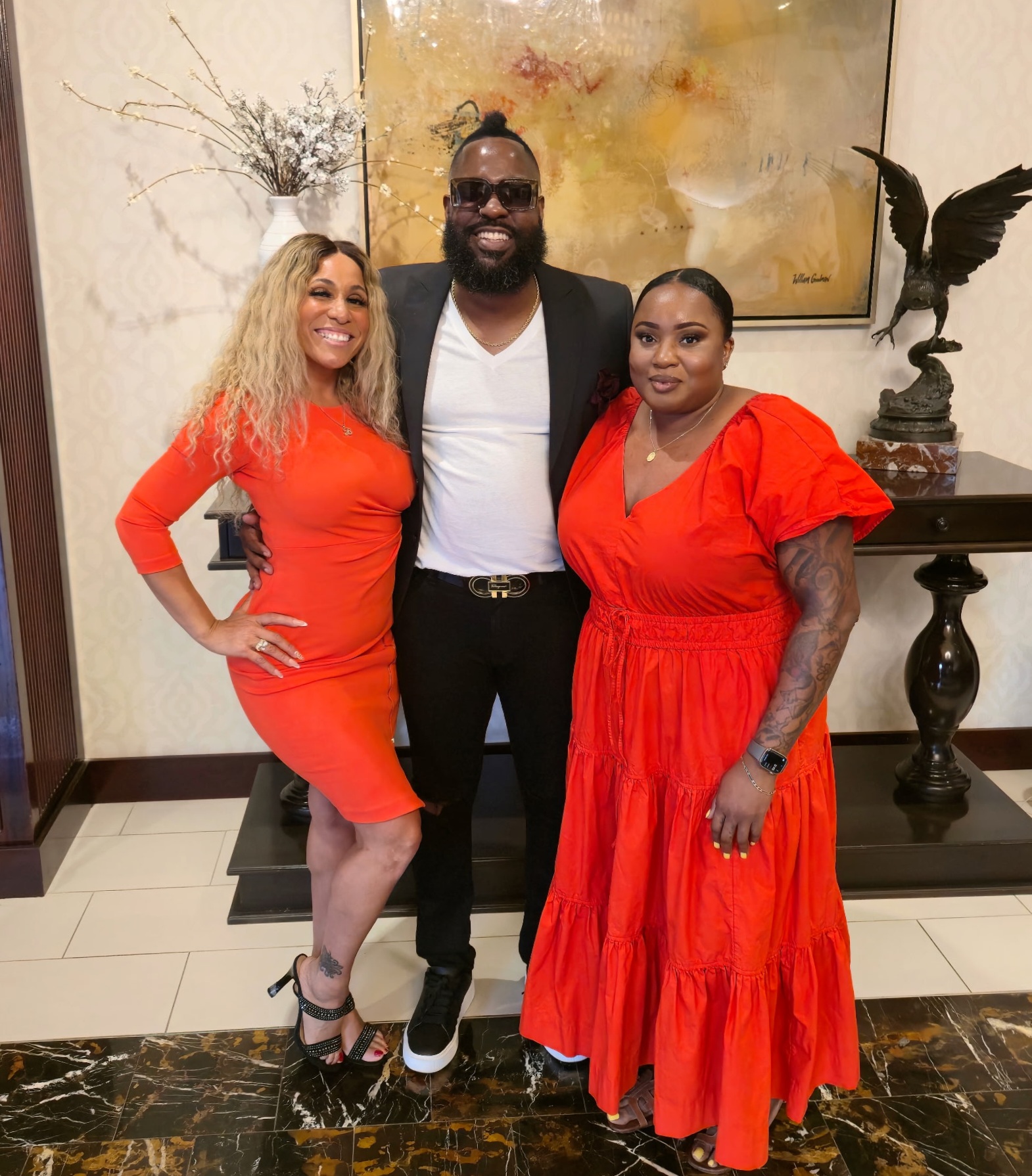 Three people standing indoors, two women in red dresses and a man in a black suit, smiling for the photo.