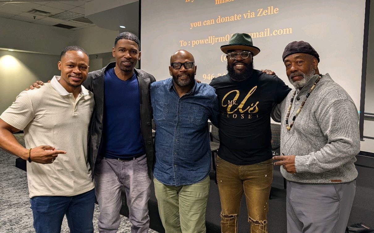Five men stand together indoors, smiling, with a screen displaying text behind them.