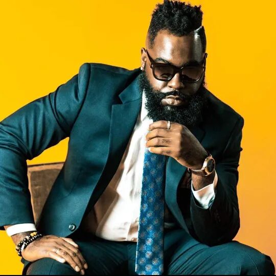 Man in suit with sunglasses, sitting against yellow background, resting chin on hand, wearing watch and bracelets.