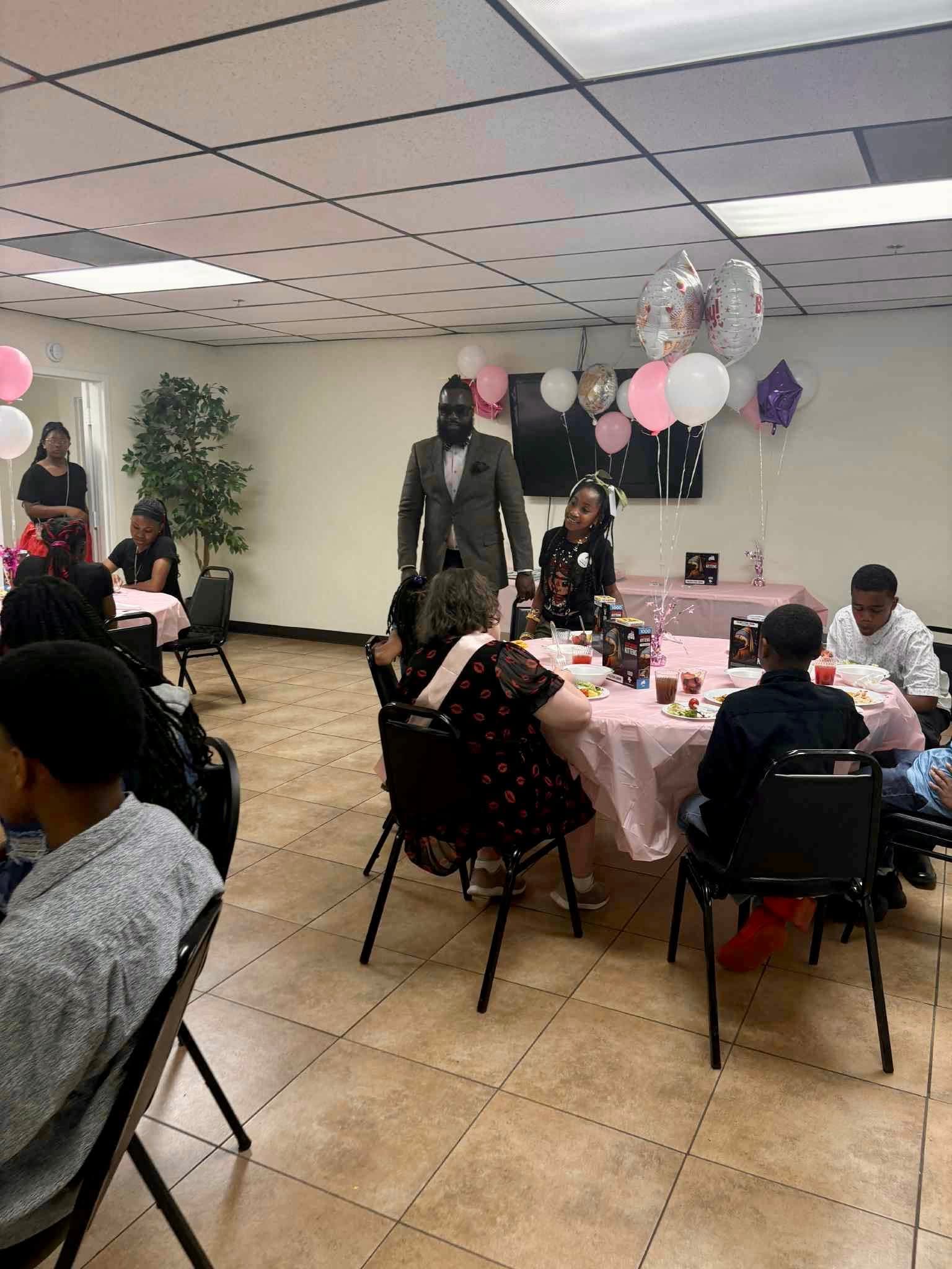 People gathered around a decorated table with balloons, a mannequin, and a plant in a room with tiled floor and ceiling tiles.