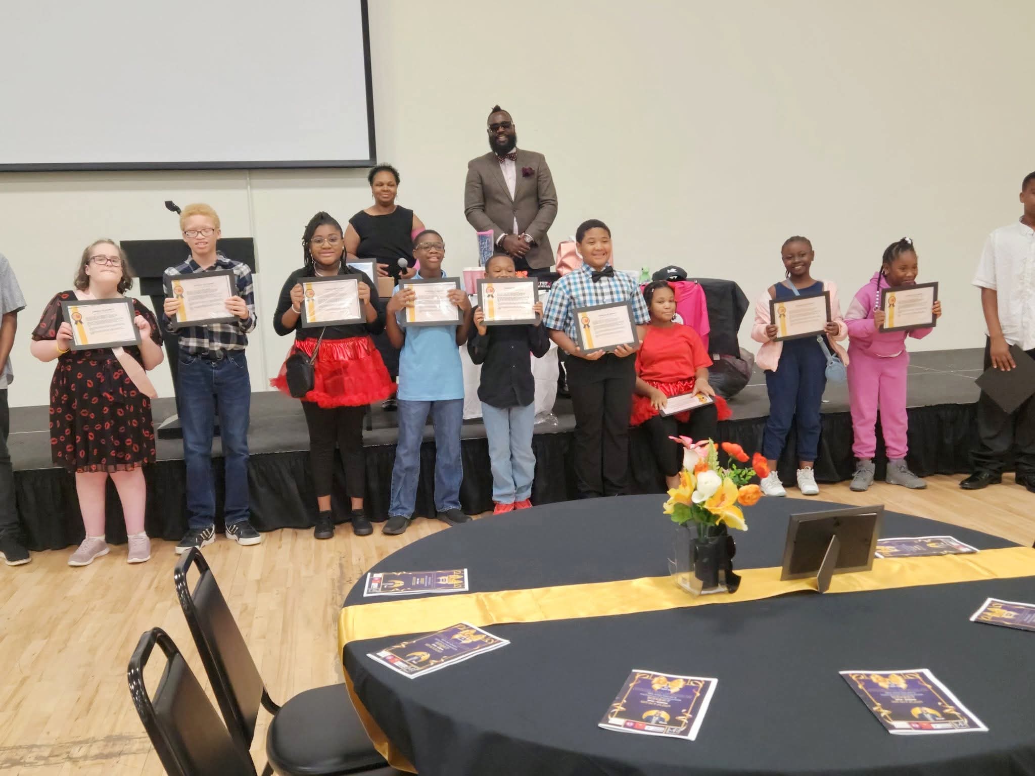 Group of children holding certificates on stage with adult standing behind them, in a room with tables and flowers.